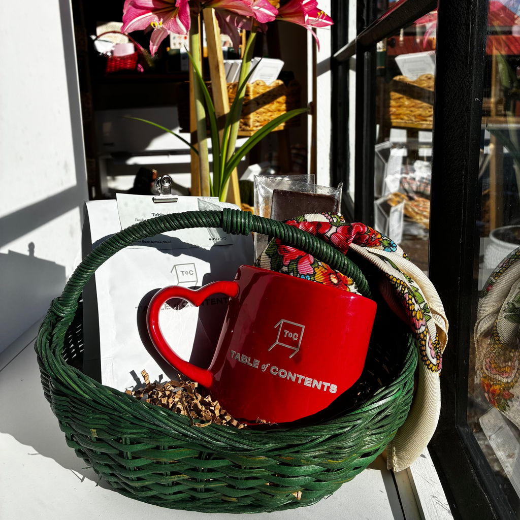 Red mug with floral arrangement and decorative items in a green basket on a table.